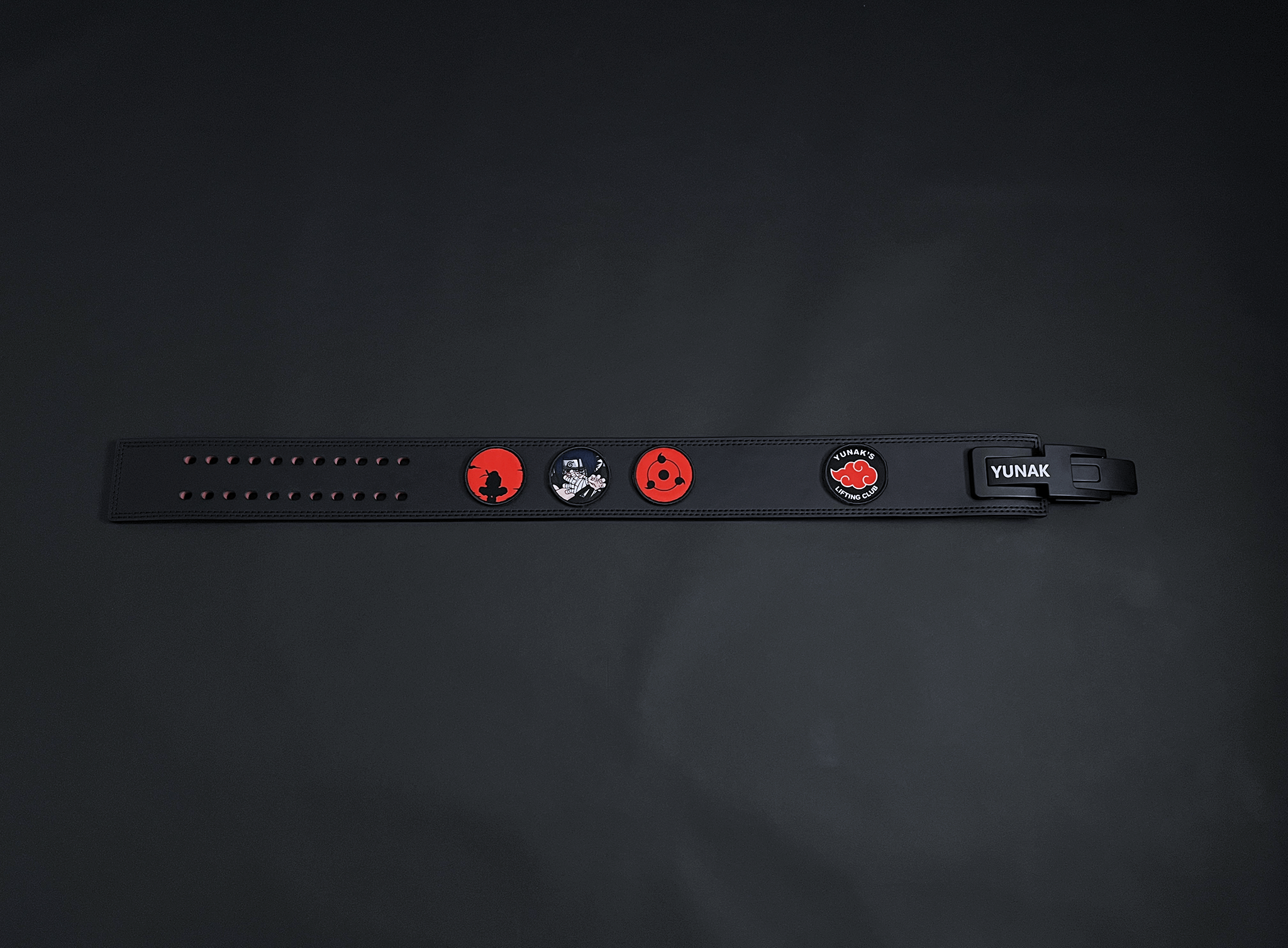 An anime powerlifting belt, 13mm thick and 4 inch wide. Featuring 4 embroidery patches inspired by the Akatsuki clan. First patch is Itachi bright red background, second is Sasuke, third is featuring the Sharingan and the fourth is a Yunak's Lifting Club patch. The photo is top down view of the belt.