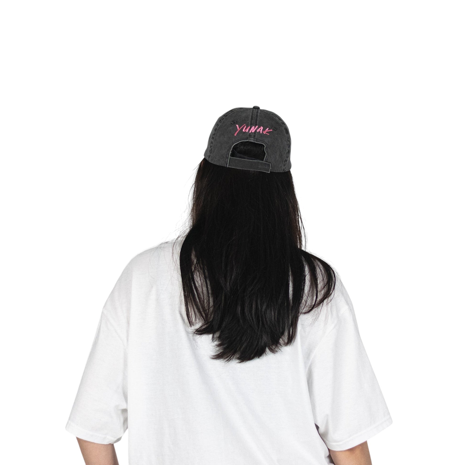 Person wearing a black vintage unstructured dad hat with embroidered pink design of Yunak on the back, standing against a white background