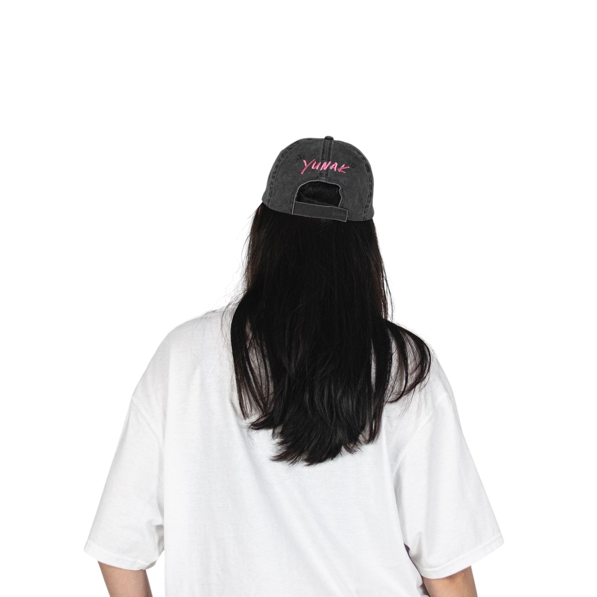 Person wearing a black vintage unstructured dad hat with embroidered pink design of Yunak on the back, standing against a white background