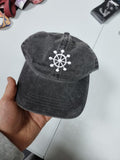 Person wearing a black vintage unstructured dad hat with embroidered white design of Mahoraga's symbol from Jujutsu Kaisen on the front, squatting against a white background