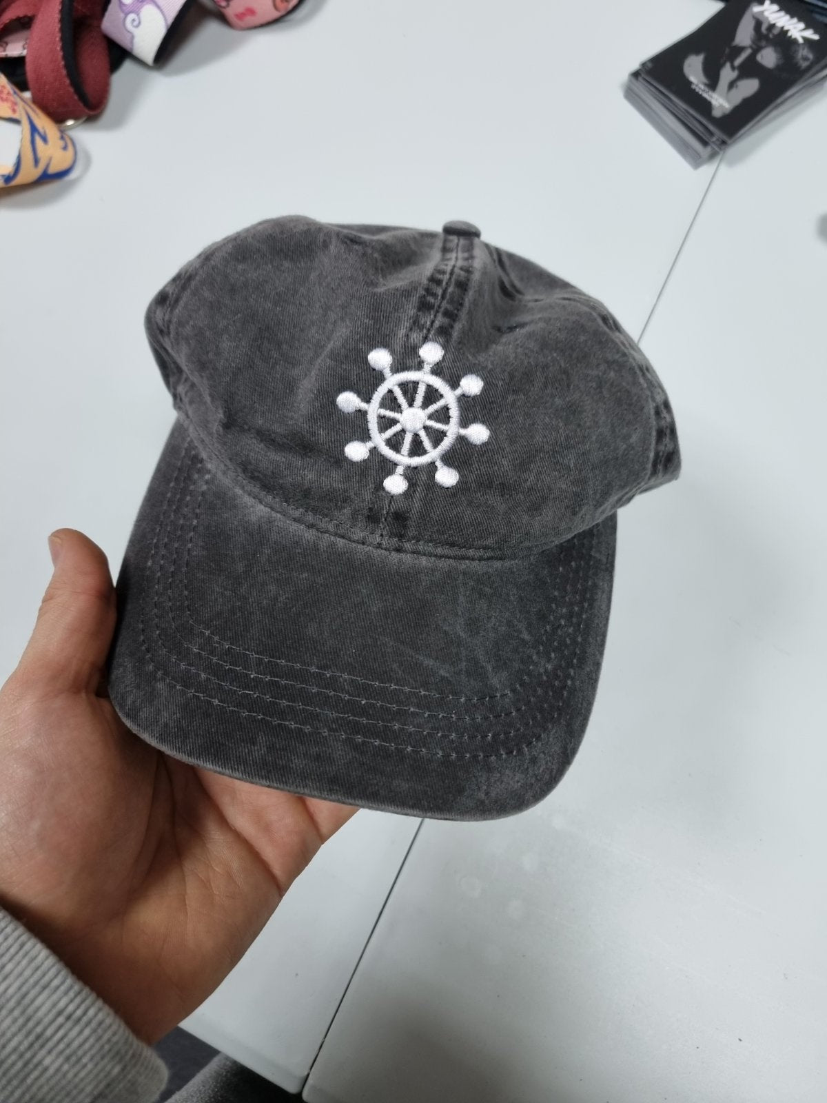 Person wearing a black vintage unstructured dad hat with embroidered white design of Mahoraga's symbol from Jujutsu Kaisen on the front, squatting against a white background
